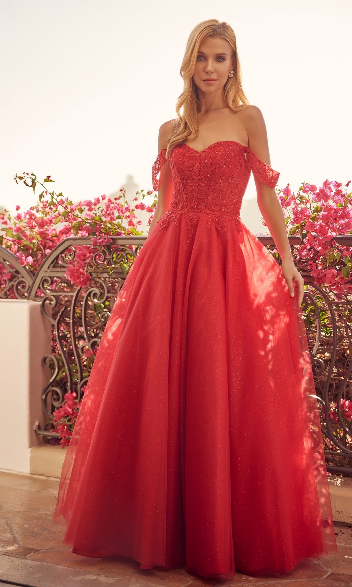 A woman stands on a balcony in the Lace-Up Off-Shoulder Long A-Line Prom Dress 280, featuring a lace corset bodice and flowing tulle skirt; pink flowers and ornate railing enhance the elegant setting.